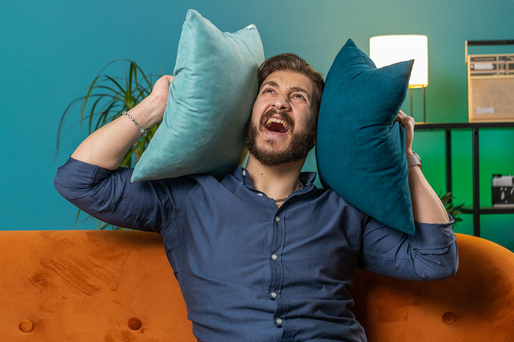 Irritated mad man relaxing on couch cover ears with pillows annoyed by noisy neighbors